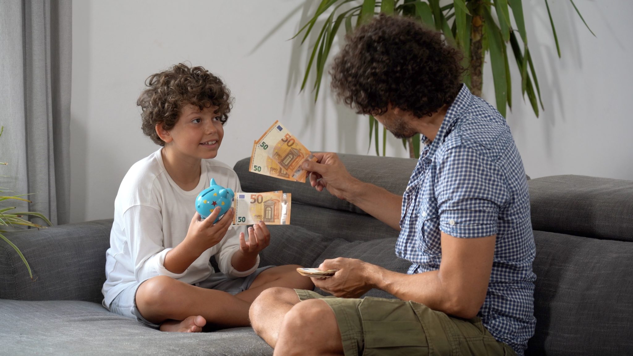 Father and son boy 9 years old    - sitting on sofa at home ion living room - the father  gives tip money for the summer holidays to the son who counts it ,  lifestyle during childhood