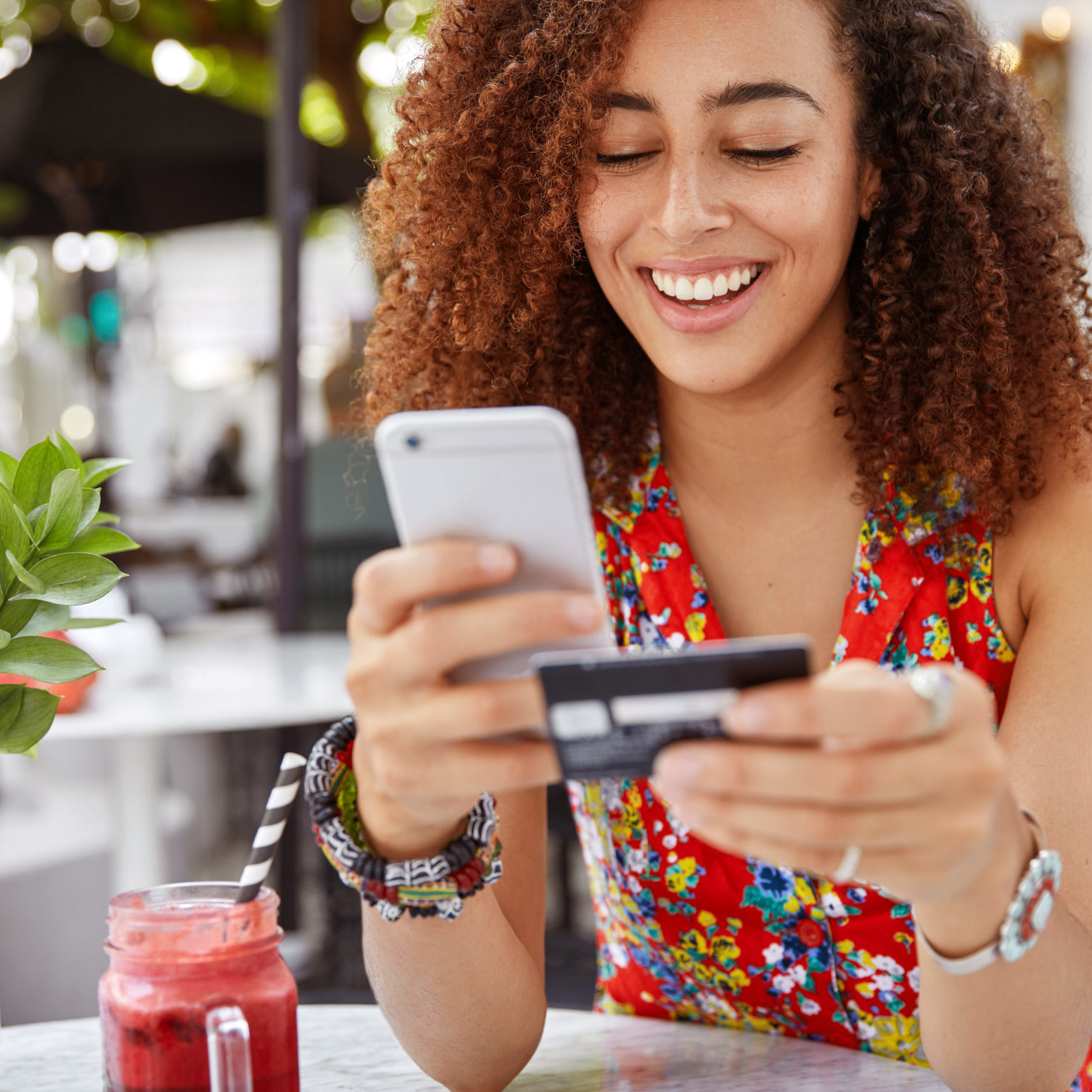 Beautiful dark skinned young female with cheerful expression, holds smart phone and credit card, banks online or makes shopping while sits against cafe interior. Payment and leisure concept.